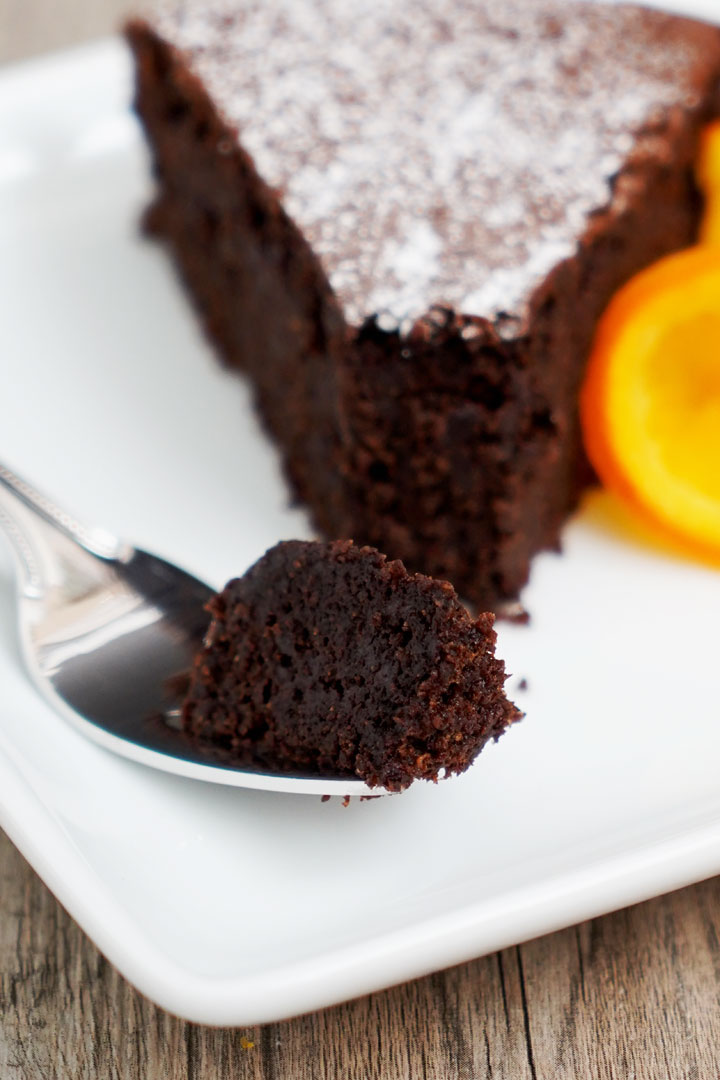 Close up view of orange and chocolate flourless cake bite on a fork with slice in background.