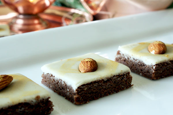 Three chocolate burfi pieces topped with a layer of marzipan and an almond on a white plate.