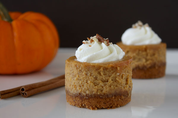 Two mini bourbon pumpkin cheesecakes on a white platter with cinnamon sticks and a pumpkin off to the side.