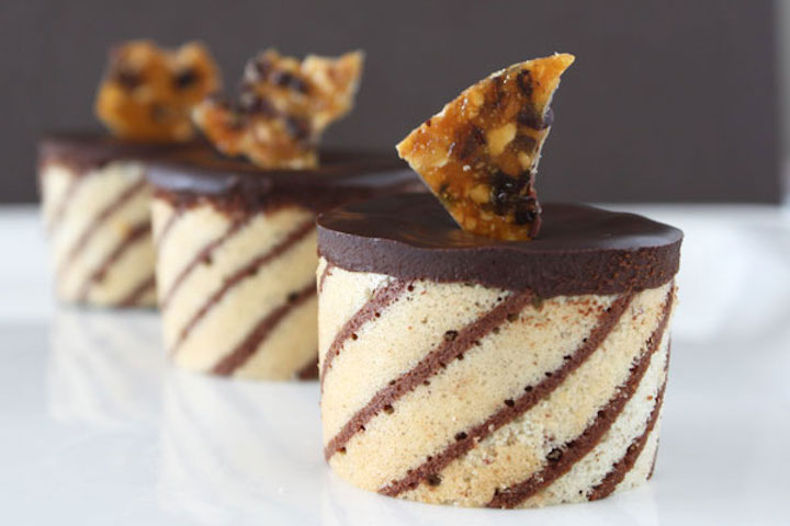 Three hazelnut mousse entremets wrapped in striped joconde and topped with hazelnut brittle, lined up diagonally on a white plate.