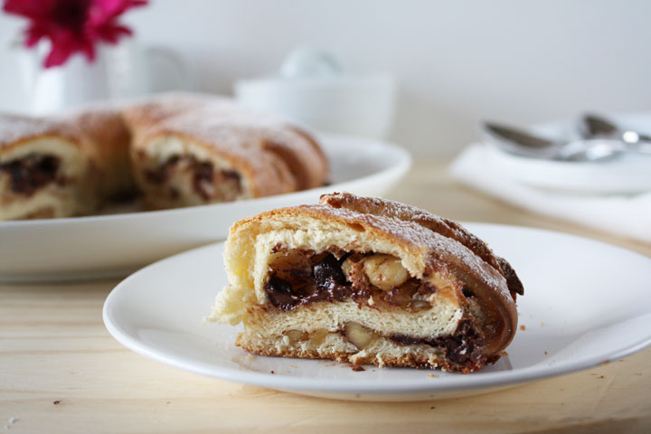 Slice of meringue Coffee Cake is stuffed with chocolate, cinnamon and walnuts on a white plate with rest of cake in the background.