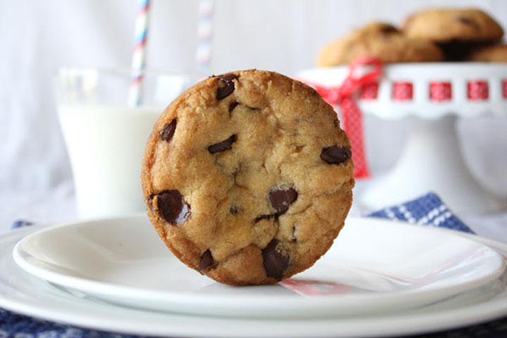 Perfectly round chocolate chip cookie standing up on a white plate with milk and cookies on cake stand in the background.