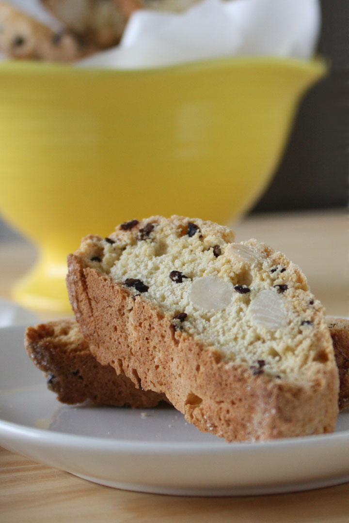 Almond biscotti with cocoa nibs on a white plate with more biscotti in a yellow bowl in the background.