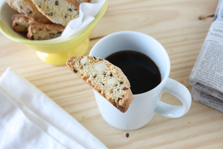 Almond biscotti with cocoa nibs sitting on top of coffee mug with more biscotti in yellow bowl and newspaper on the side.