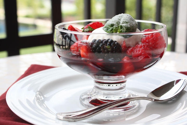 Summer berry salad cocktail with vanilla ice cream and frosted basil leaf in a clear dessert bowl in a white plate with a spoon on the side. 