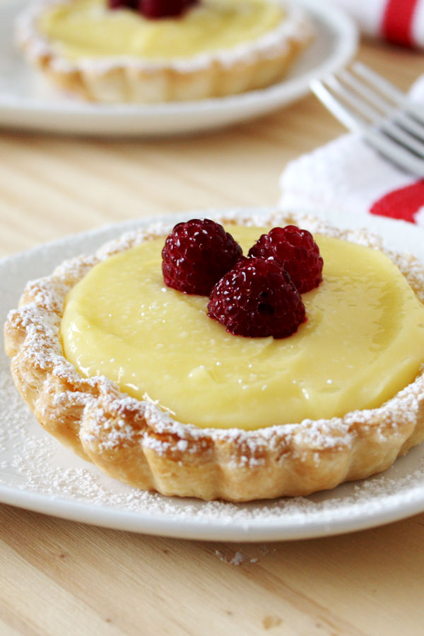Lemon Tart topped with three raspberries on a white plate with another tart and fork in the background.