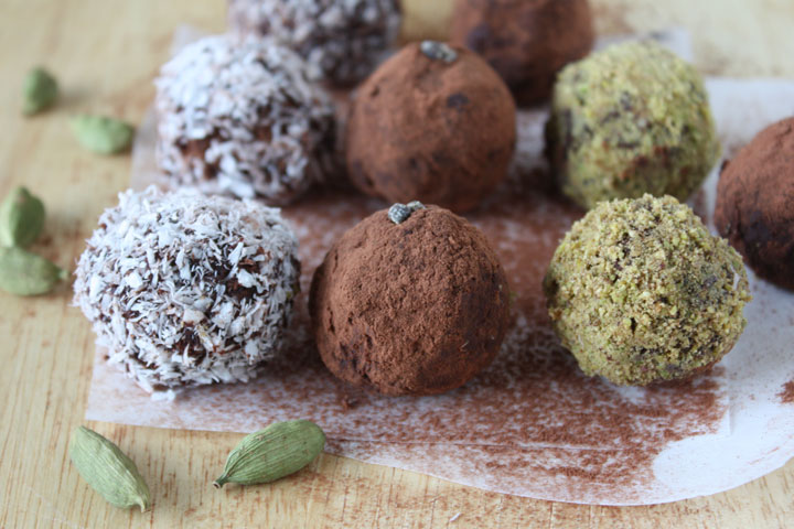 Variety of coated cardamom chocolate truffles on parchment paper with cardamom pods on side. Pistachio, Cocoa Powder and Coconut coated cardamom truffles. 