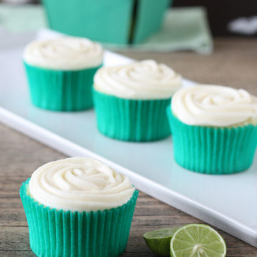 Key lime cupcake with cream cheese frosting in green cupcake wrapper with cupcakes in background and key limes on the side.