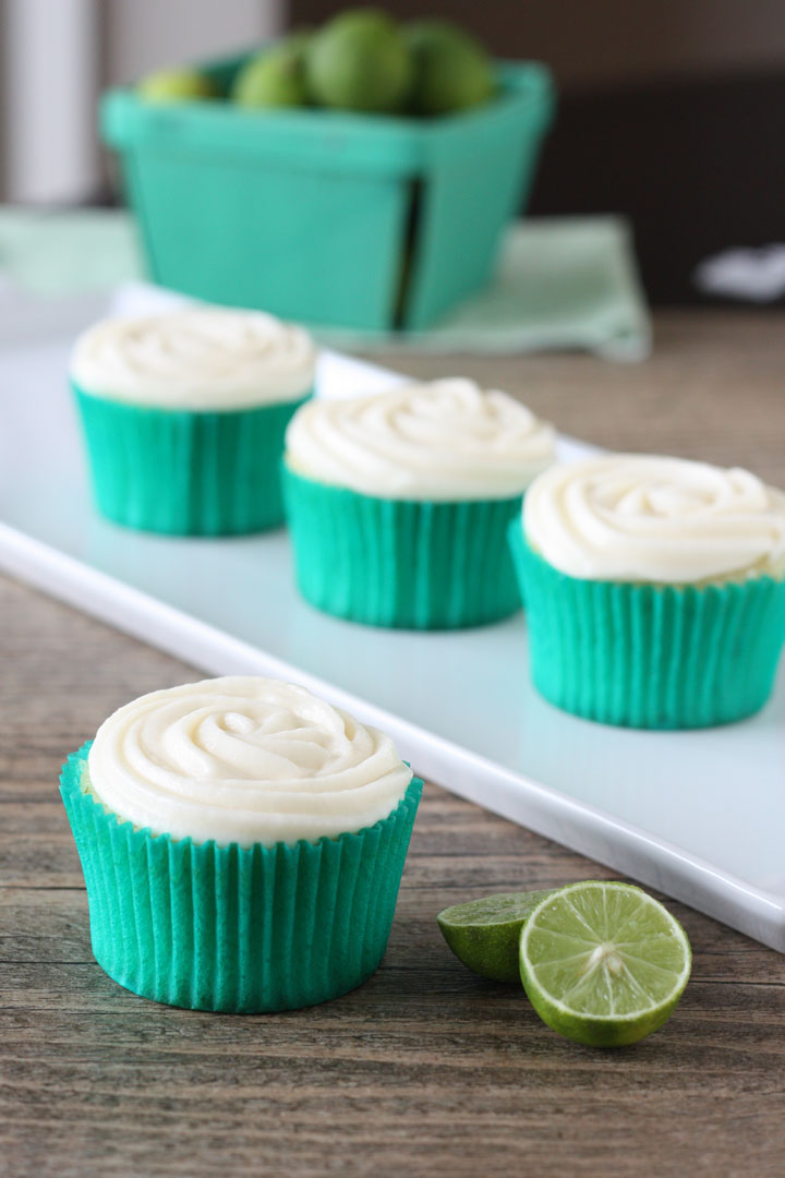 Key lime cupcake with cream cheese frosting in green cupcake wrapper with cupcakes in background and key limes on the side.