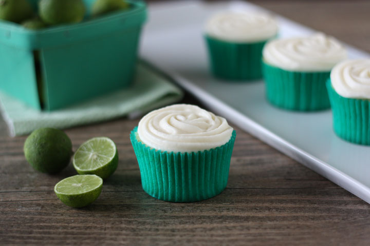 Key lime cupcake with cream cheese frosting in green cupcake wrapper with cupcakes in background and key limes on the side.