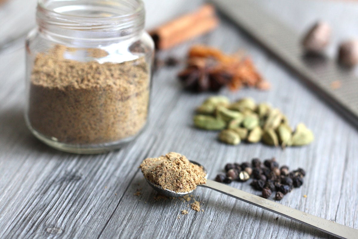 Blended chai spice mix in a spoon with jar and whole spices in background.
