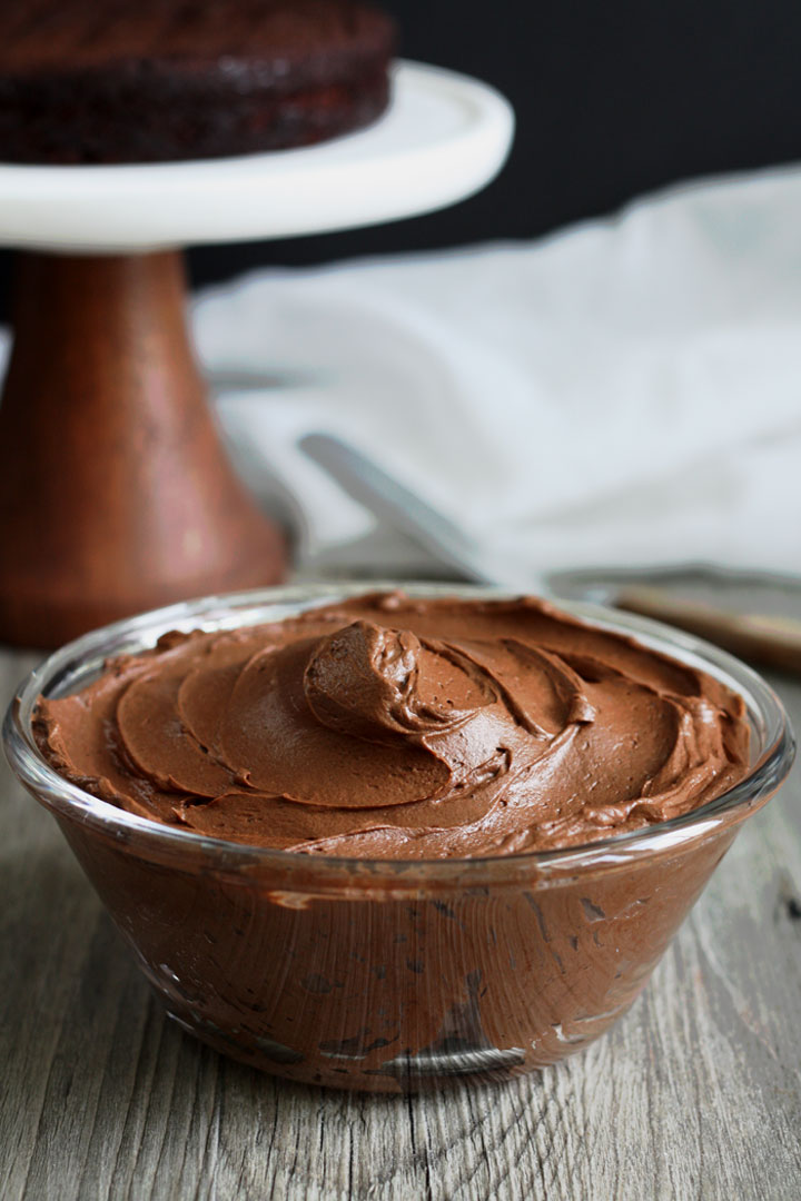 Chocolate fudge frosting in a clear glass bowl with chocolate cake on stand in the background.