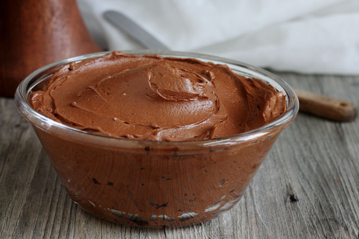 Chocolate fudge frosting in a clear glass bowl with spatula in background.