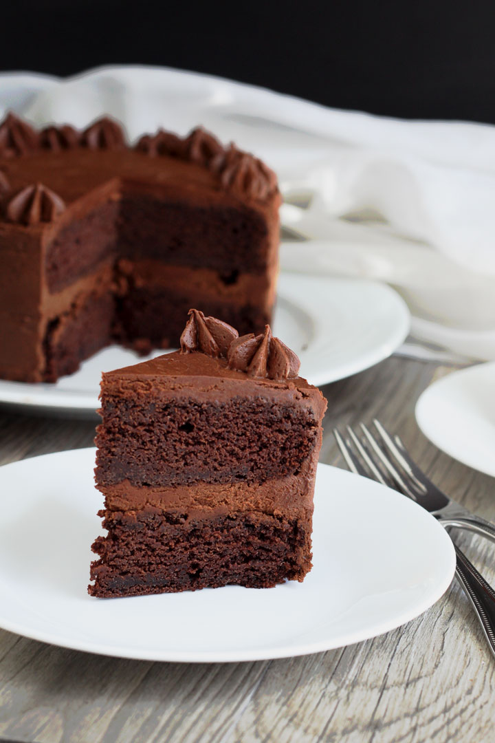 Slice of chocolate cake with fudge frosting in white plate with whole cake in background.