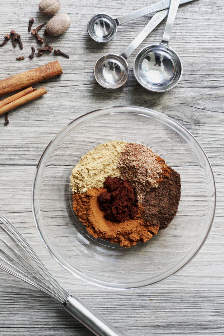 Pumpkin spice ground ingredients in a glass bowl with measuring spoons, whisk and whole ingredients on the side. 