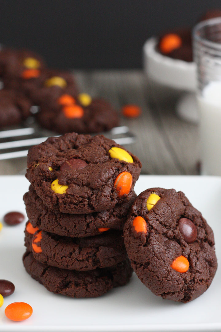 Reese's Pieces double chocolate cookies stacked in a white plate with milk and cookies in background.