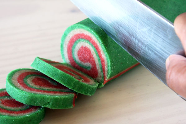 A log of unbaked pinwheel christmas nankhatai cookies being sliced with a large knife.