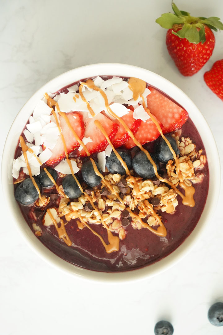 Top vie of Acai bowl topped with coconut flakes, strawberries, blueberries, granola, chocolate chips and peanut butter drizzle.