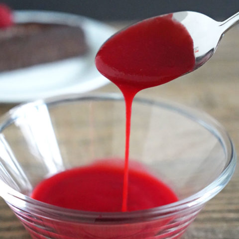 Raspberry Coulis being poured off a spoon into a bowl full of more coulis. Slice of chocolate cake is in the background.