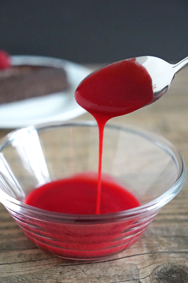 Raspberry Coulis being poured off a spoon into a bowl full of more coulis. Slice of chocolate cake is in the background.