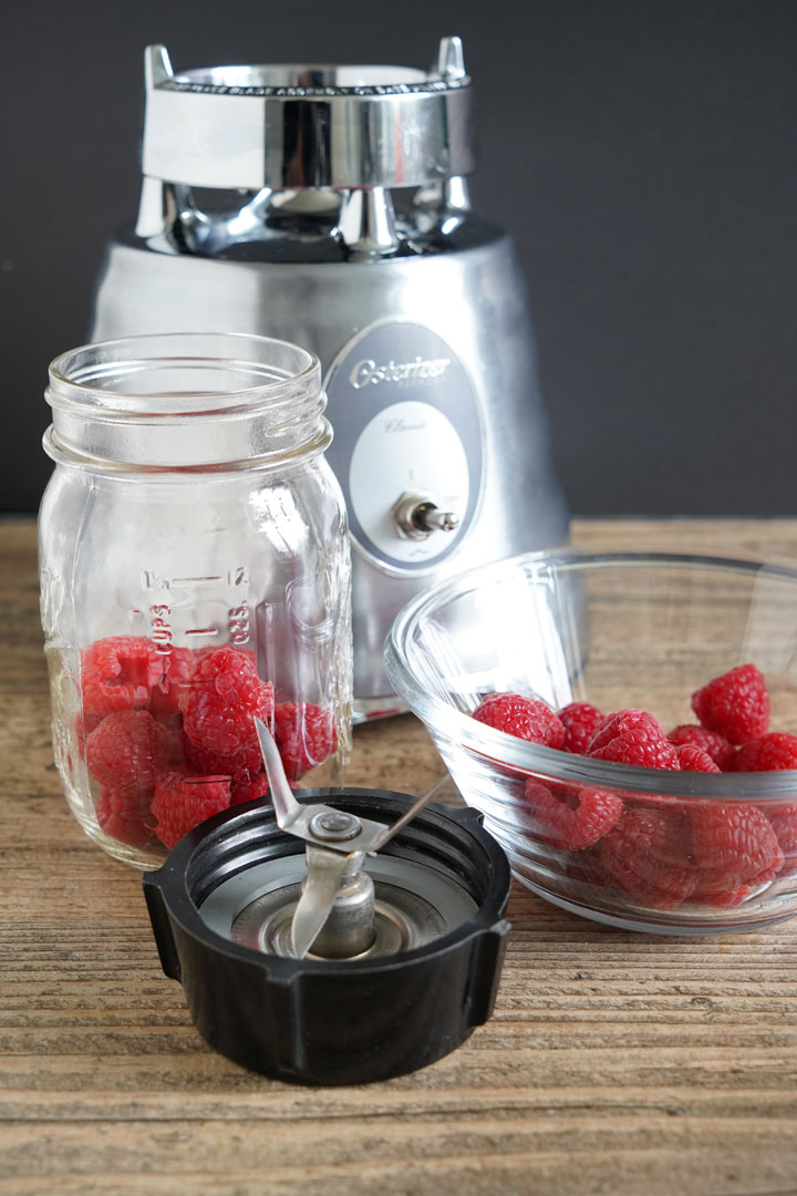 Fresh raspberries in a mason jar next to a bowl of more raspberries, a blender base, and a blender blade.