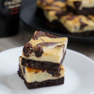 Two Irish cream cheesecake brownies on a white plate with Baileys bottle and more brownies on a black plate in the background.