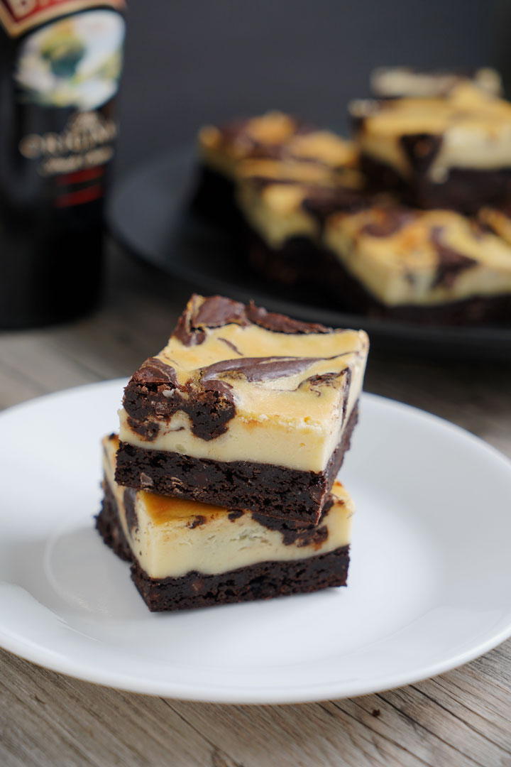 Two Irish cream cheesecake brownies stacked on white plate with Baileys bottle and more brownies on a plate in the background and m
