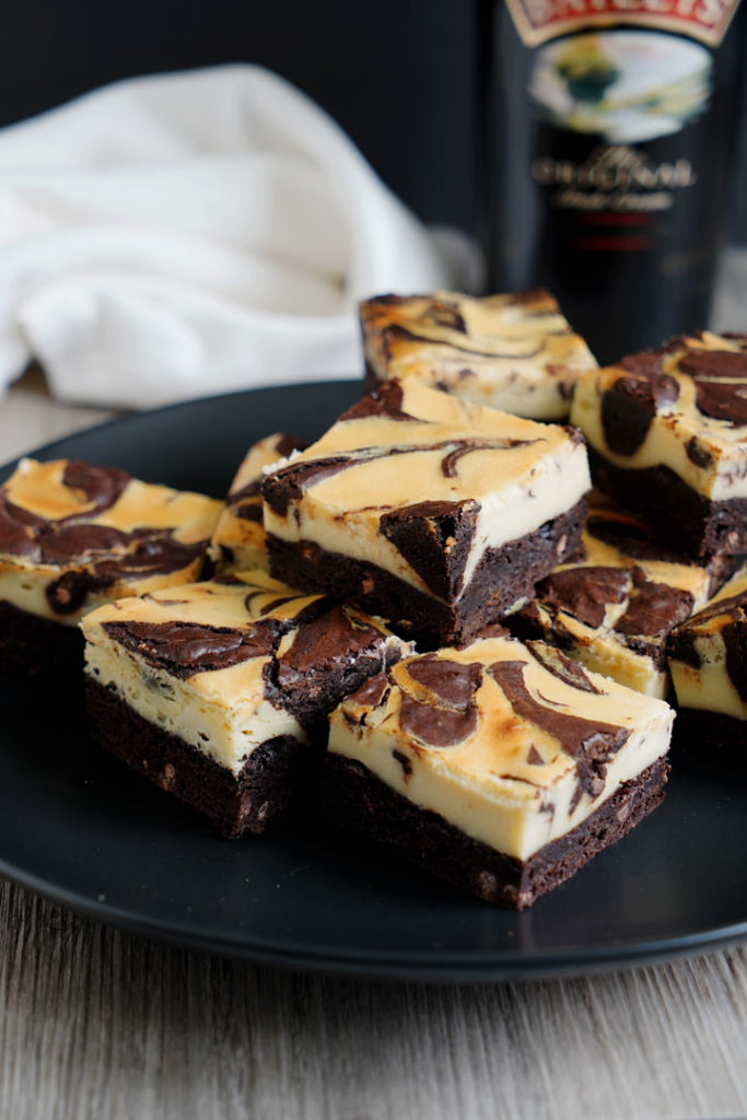 Irish cream cheesecake brownies on a black dish with Baileys bottle in background.