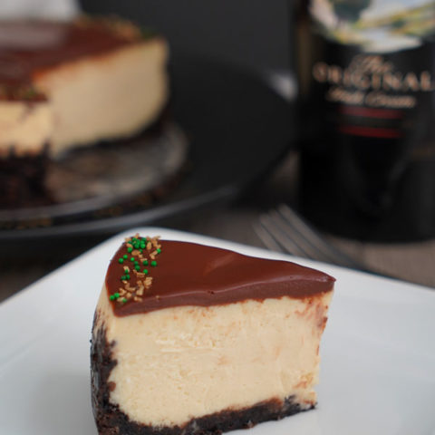 Slice of Baileys Cheesecake on white plate with cheesecake and bottle in background.