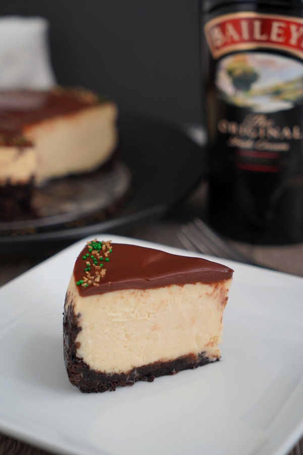 Slice of Baileys Cheesecake on white plate with cheesecake and bottle in background.