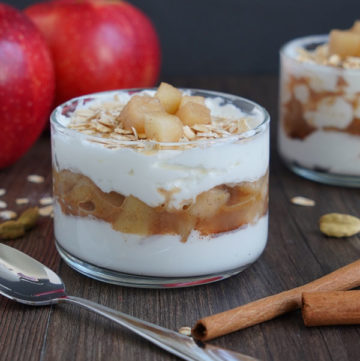 Cooked Apple parfait in small glass bowl with spoon on the side. Apples in background and cinnamon sticks in the foreground.