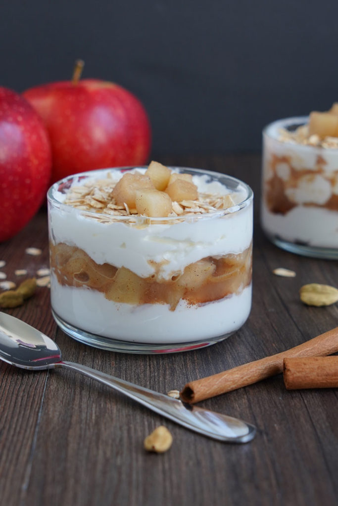 Apple Cinnamon Parfait in small glass bowl with spoon on the side. Apples in the background and cinnamon sticks in the foreground.