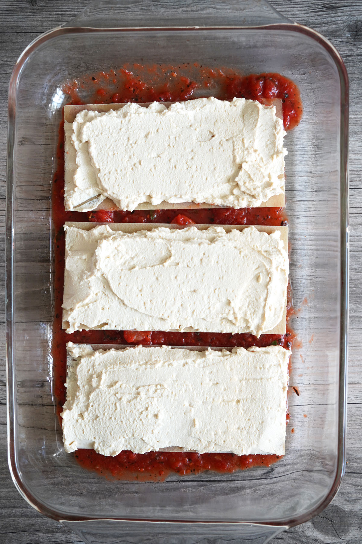 Lasagna partially assembled in glass baking dish with one layer of noodle topped with tofu ricotta. 