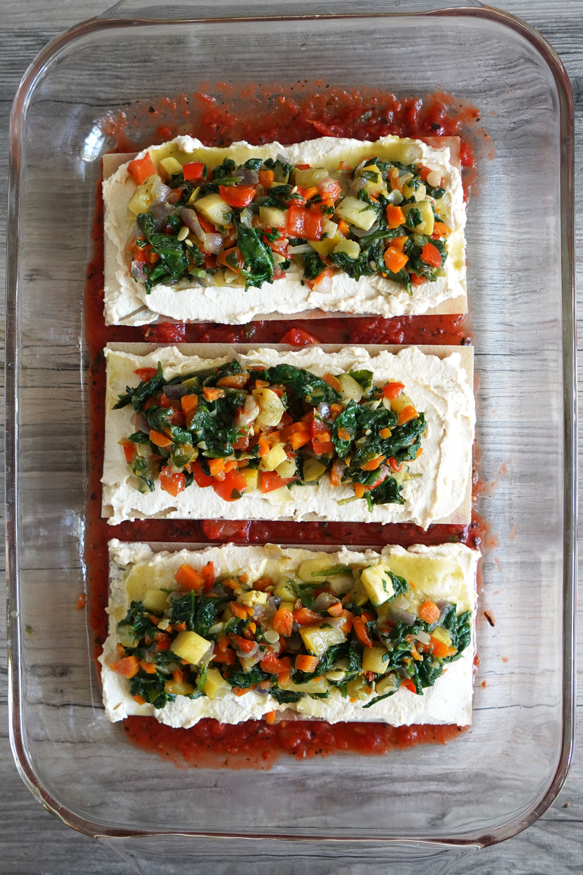 Lasagna partially assembled in glass baking dish with one layer of noodle topped with tofu ricotta and vegetables.