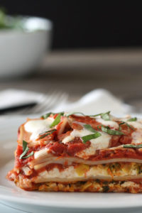 Slice of vegetable lasagna in a white plate with greens in a bowl in background.