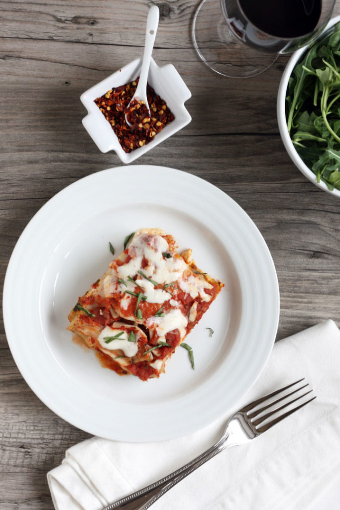 Overhead shot of vegetable lasagna slice, crushed red peppers in small dish and fork on napkin.