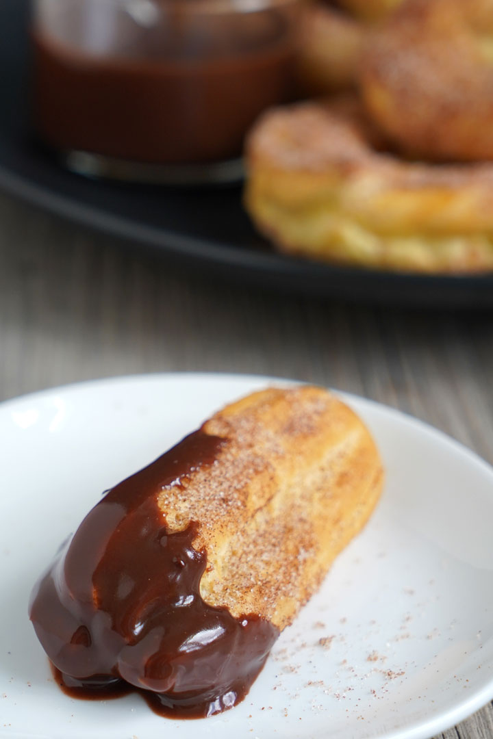 Baked churro dipped in chocolate sauce on a white plate with more churros and sauce in the background. 