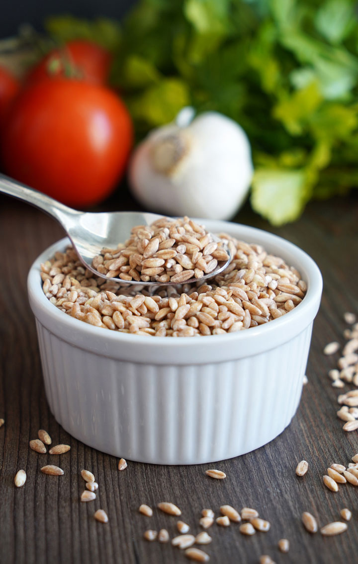 Uncooked farro in white bowl with spoon in farro and vegetables in the background.