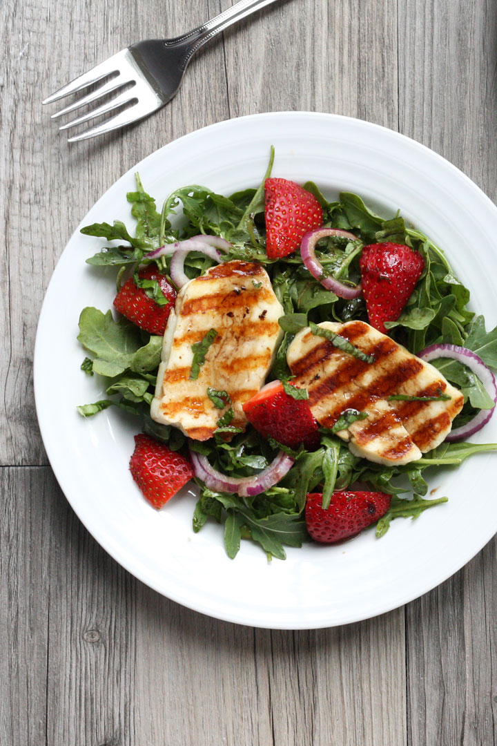 Grilled halloumi slices over a bed of arugula and strawberry salad on a white plate.