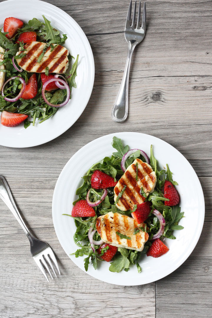 Top view- Grilled halloumi slices over a bed of arugula and strawberry salad on a white plate.