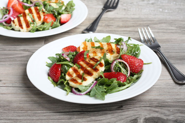 Grilled halloumi slices over a bed of arugula and strawberry salad on a white plate.