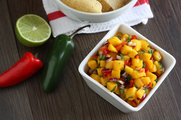 Top view Mango salsa in a square white bowl with jalapenos and lime on the side.