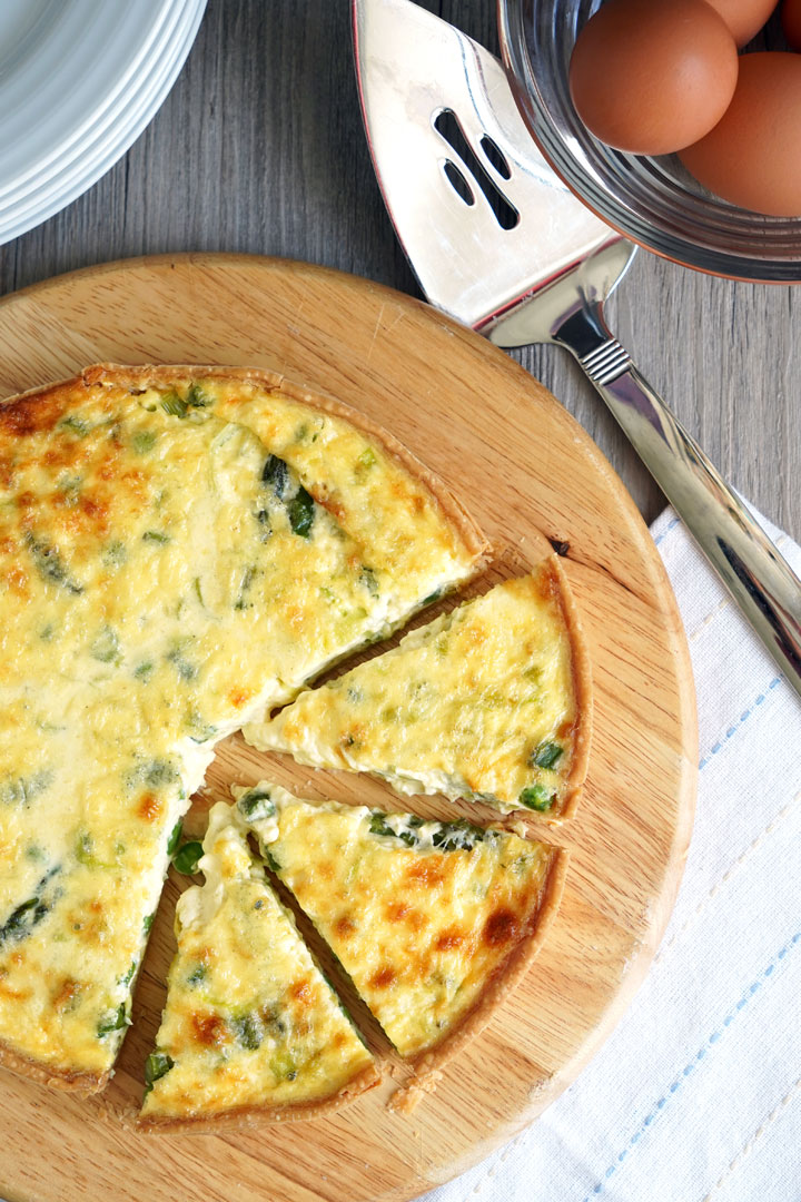 Top view of vegetable quiche on a cutting board with eggs and plates on the side.