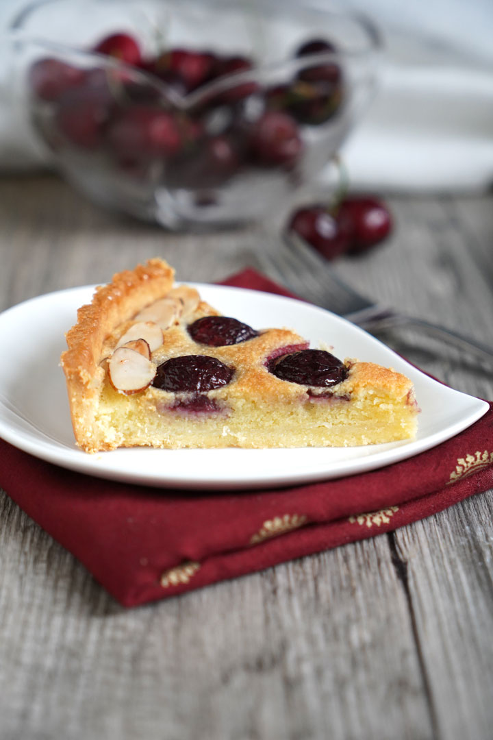 Slice of cherry tart with almond cream filling on a white plate and a red napkin with cherry bowl in the background. 