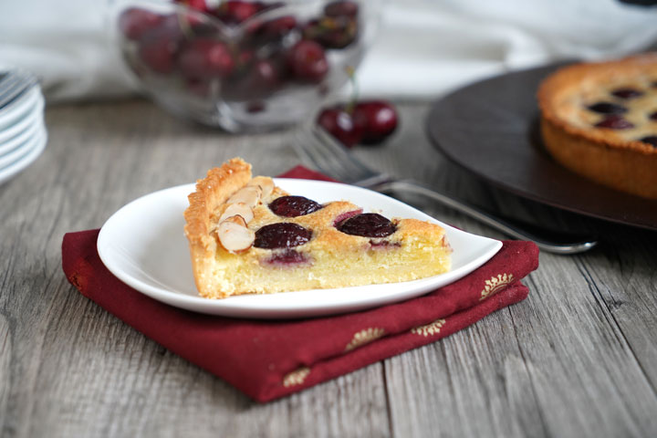 Slice of cherry tart with almond cream filling on a white plate and a red napkin with cherry bowl and tart in the background. 