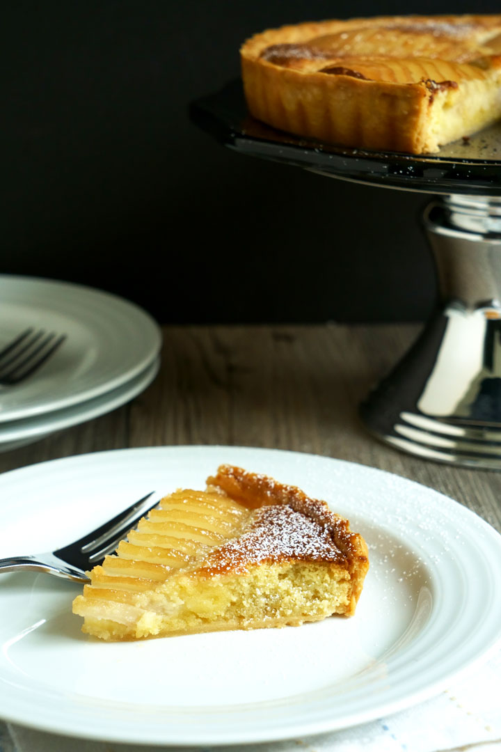 Slice of pear and frangipane tart on a white plate with fork and tart on cake stand in the background.