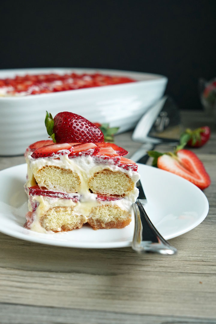 Slice of strawberry tiramisu on a white plate with fork. Strawberries and cake server in background.