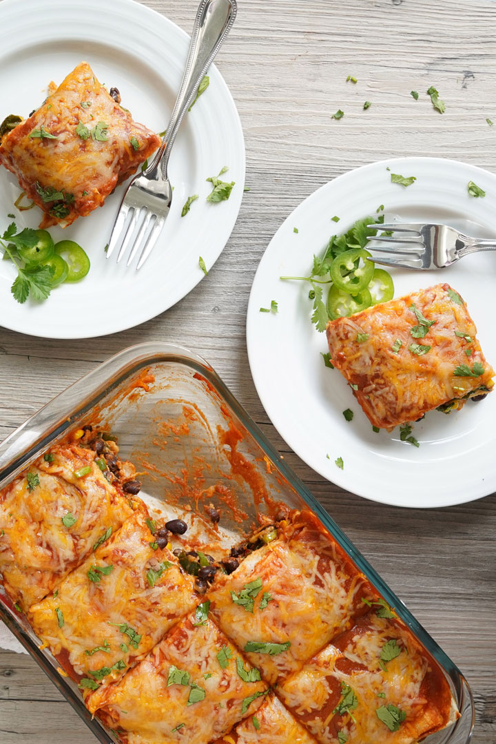 Top view of casserole tray and pieces of enchilada casserole on white plates with forks. 