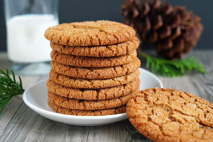 Stack of gingersnap cookies with one cookie off of plate and milk glass and pine cones in the background.