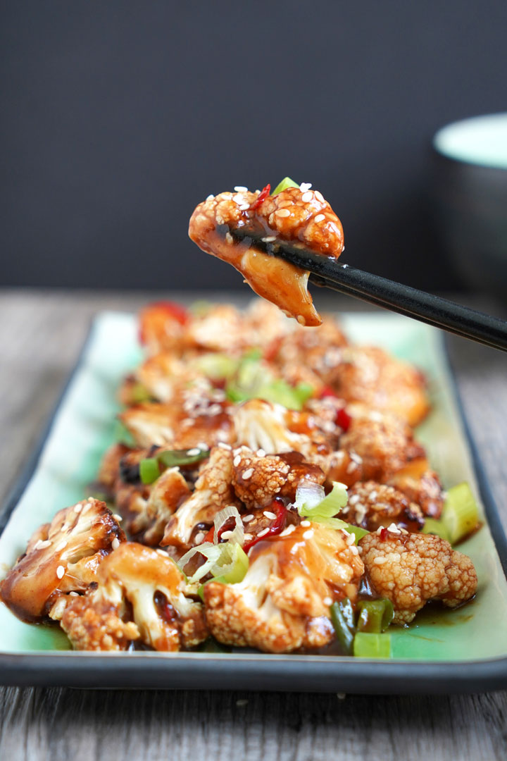 Piece of Kung Pao Cauliflower being held between chopsticks with cauliflower in plate in background.